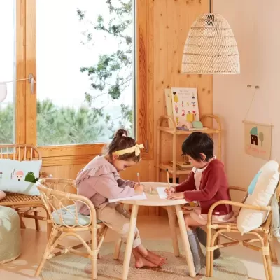 Crianças em cadeiras de vime a desenhar numa mesa redonda de madeira clara, com janela grande e candeeiro de fibras naturais.