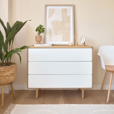 Cómoda de madeira clara com gavetas brancas, moldura, plantas e cadeira branca