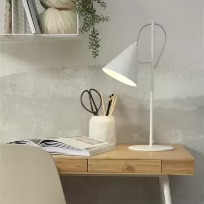 Secretária de madeira clara com lâmpada branca, jarro com tesoura e lápis, livro aberto e cadeira creme num ambiente minimalista.