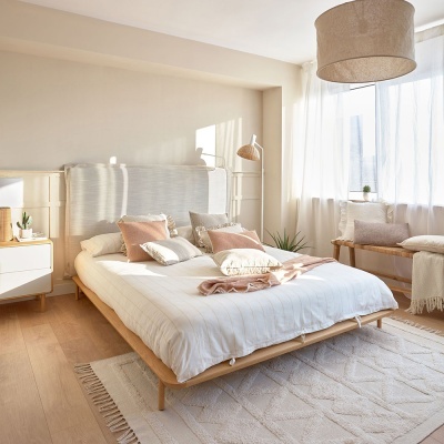 Quarto com cama de madeira clara, roupa de cama branca, almofadas bege e rosa, candeeiro dourado e cortinas brancas