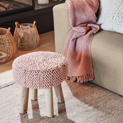 Pufe redondo com assento em malha rosa e pernas de madeira na sala, ao lado de sofá com manta rosa e cestos em bambu