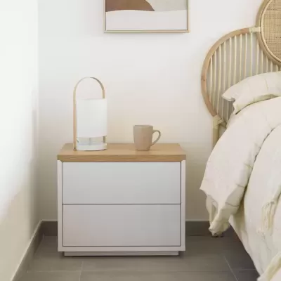 Mesa de cabeceira branca com tampo em madeira clara, lâmpada e caneca junto a cama com cabeceira de rattan