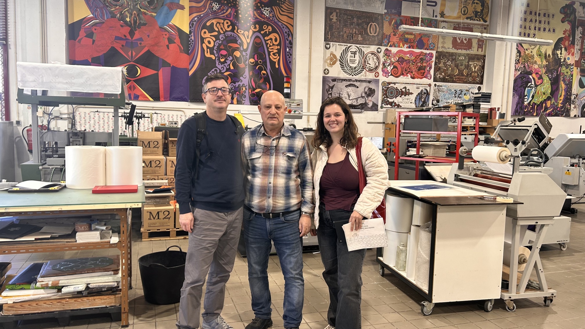 Três pessoas numa oficina com máquinas industriais e obras de arte nas paredes