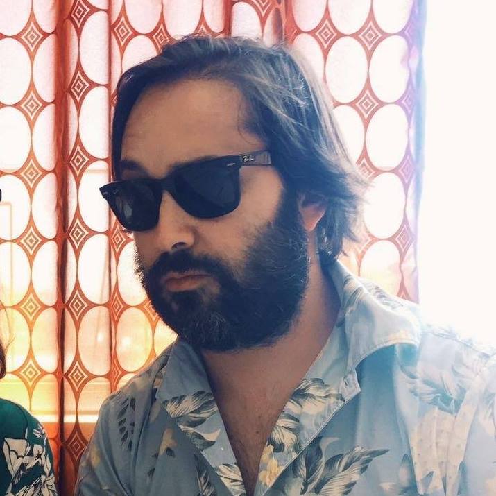 Homem com óculos de sol pretos e camisa azul floral em frente a cortina vermelha