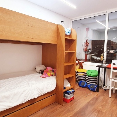 Quarto com cama de madeira clara tipo beliche, mesa e cadeira brancas, armário escuro e janela grande
