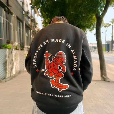 Camisola preta de streetwear com estampa vermelha e branca nas costas com personagem de skate e texto, ambiente urbano com árvores