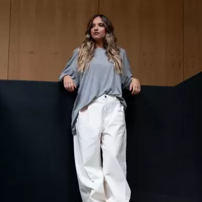 Mulher com t-shirt cinzenta e calças brancas encostada em estrutura preta