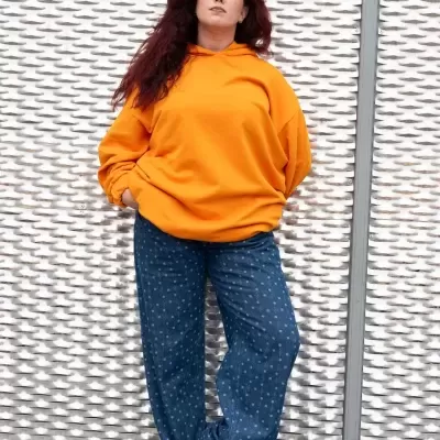 Mulher com hoodie laranja e calças azuis de pontos brancos junto a fundo metálico