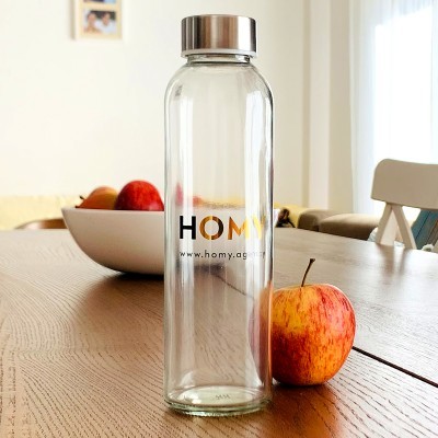 Garrafa de vidro transparente com tampa metálica e logo HOMY em mesa de madeira junto a maçã.