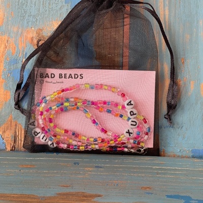 Colar de contas coloridas com palavras em letras brancas, saco de organza preto e cartão rosa sobre madeira azul e castanha.