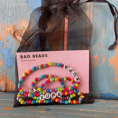 Conjunto de pulseiras coloridas com contas e letras 'XUPA' em saco preto de organza com cartão Bad Beads