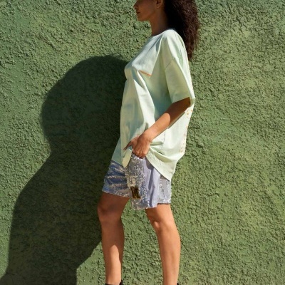 Mulher com t-shirt verde clara, saia cinza com padrão e sandálias pretas de salto junto a parede verde