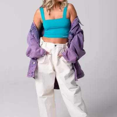 Mulher com top azul, calças brancas largas e casaco roxo oversized, fundo cinzento.