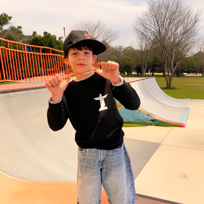 Criança com camisola preta, calções de ganga, boné preto, colar dourado, em skatepark com guarda-corpo laranja.