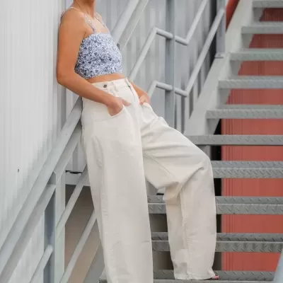 Mulher com calças largas brancas e top azul com padrão floral, óculos de sol, encostada em parede metálica junto a escada metálica cinza.