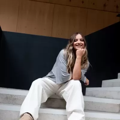 Mulher sorridente sentada numa escadaria, roupa casual clara, fundo escuro e madeira