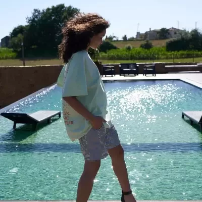 Mulher perto de piscina, camisa azul, calções cinza e sandálias pretas de salto alto