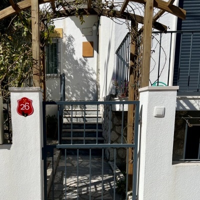 Portão cinza em muro branco com placa número 26 e arco de madeira com plantas secas