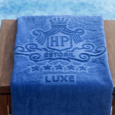 Toalha azul de algodão com relevo 'HP ESTORIL LUXE' sobre banco de madeira junto a piscina