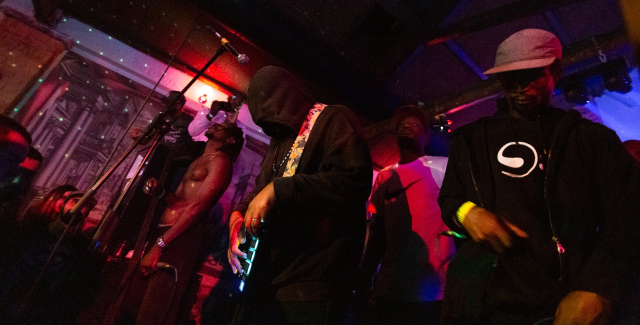 Quatro homens numa sessão musical em palco escuro com luzes vermelha e azul.