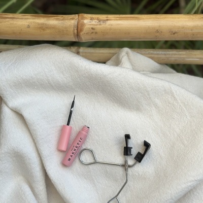 puxador de pestanas metálico e máscara de pestanas rosa sobre tecido branco com bambu e plantas ao fundo