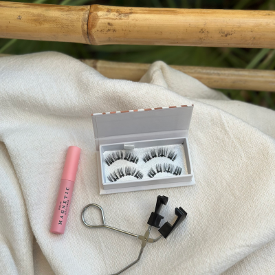 Kit de produtos para pestanas com tubo rosa 'MAGNETIC', pestanas falsas em caixa branca e aplicador metálico posicionado sobre tecido branco e bambu no fundo