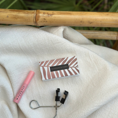 Conjunto de modelador de pestanas, batom rosa e caixa de pestanas de padrão listrado sobre tecido bege junto a bambu.