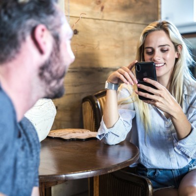 Duas pessoas numa mesa de madeira interior, mulher loira com smartphone.
