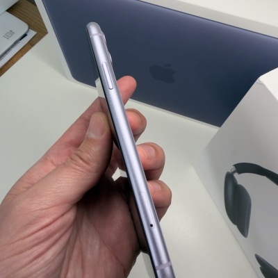 Smartphone cinza metálico segurado em mão com caixas de MacBook NEO e auscultadores ao fundo.