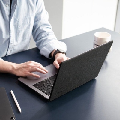 Portátil cinza escuro com textura tipo tecido em mesa azul, pessoa a usar, caneta digital branca e caneca branca ao lado