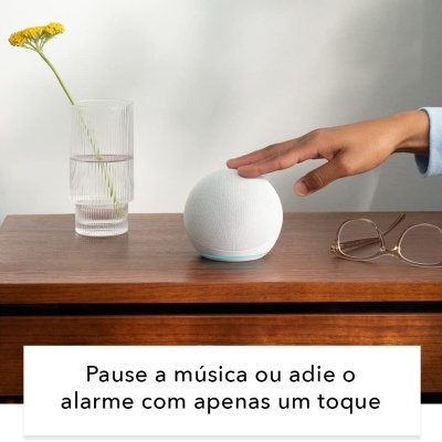 Dispositivo esférico branco sobre mesa com copo de água e flor, óculos e mão prestes a tocar dispositivo