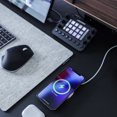 Estação de trabalho com rato, teclado, smartphone a carregar, earphones e dispositivo Elgato Stream Deck numa secretária.