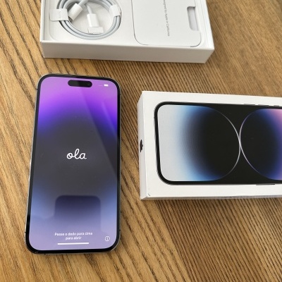 Smartphone preto com ecrã a mostrar 'ola' e caixa com cabo USB-C sobre mesa de madeira