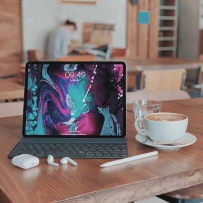 Tablet com teclado, café com leite, copo de água, caneta e auriculares sobre mesa de madeira num café