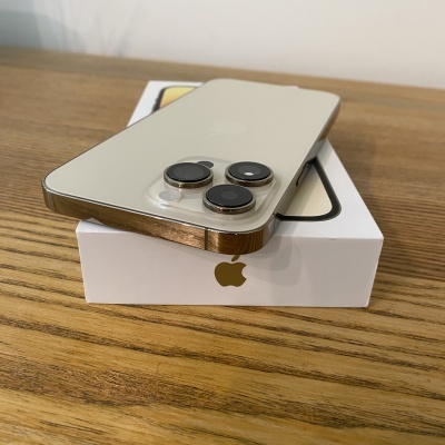 Smartphone dourado com três câmaras traseiras em cima de uma caixa branca da Apple numa mesa de madeira