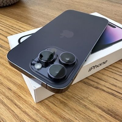 iPhone preto com três câmaras traseiras sobre caixa branca com texto 'iPhone' em mesa de madeira