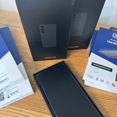 Celular Samsung Galaxy S24 preto em estojo aberto com duas caixas fechadas e folhetos sobre protetor de ecrã na mesa de madeira