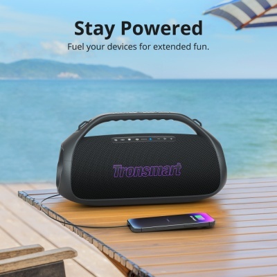 Coluna portátil preta Tronsmart com telemóvel em carregamento numa mesa junto ao mar