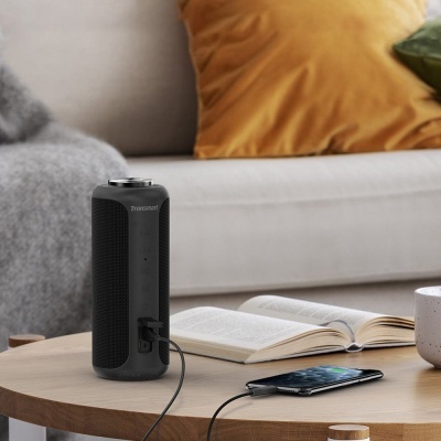 Coluna portátil preta Tronsmart numa mesa de madeira com livro aberto, telemóvel e caneca, em ambiente de sala com sofá e almofadas.
