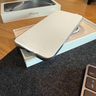 Smartphone iPhone branco sobre caixa aberta com cabo dentro, em mesa de madeira, perto de teclado e embalagem Samsung no fundo.