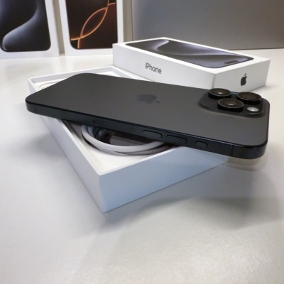 iPhone preto de lado com três câmaras sobre caixa aberta com cabo, caixas e portátil Apple ao fundo