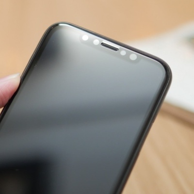 Smartphone preto com moldura preta e sensores no topo, segurado por mão, fundo com mesa clara