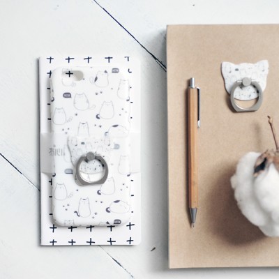 Capa de telemóvel branca com padrão de gatos e anel metálico, caneta de madeira e caderno castanho com suporte para anel