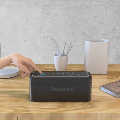Coluna de som portátil Tronsmart preta numa mesa de madeira com vasos brancos e um livro