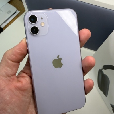 Iphone lilás com câmara dupla e logótipo Apple dourado na mão com caixa de MacBook Neo ao fundo