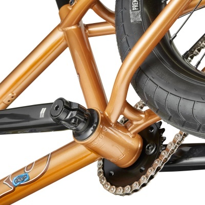 Detalhe do quadro dourado, pedal preto, corrente e pneu da bicicleta.