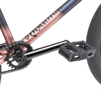 Detalhe do pedal, braço da pedaleira, corrente, roda traseira e quadro rosa metálico com texto preto de bicicleta