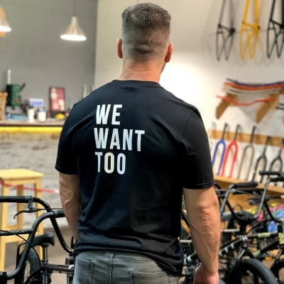 Camiseta preta com texto branco WE WANT TOO nas costas em ambiente com bicicletas