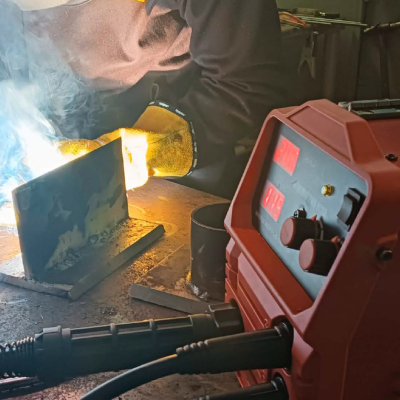 Máquina de soldar vermelha e peça metálica a ser soldada por pessoa com luvas amarelas