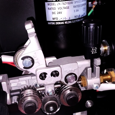 Mecanismo de alimentação de arame para soldadura preto com engrenagens metálicas e etiqueta com especificações técnicas